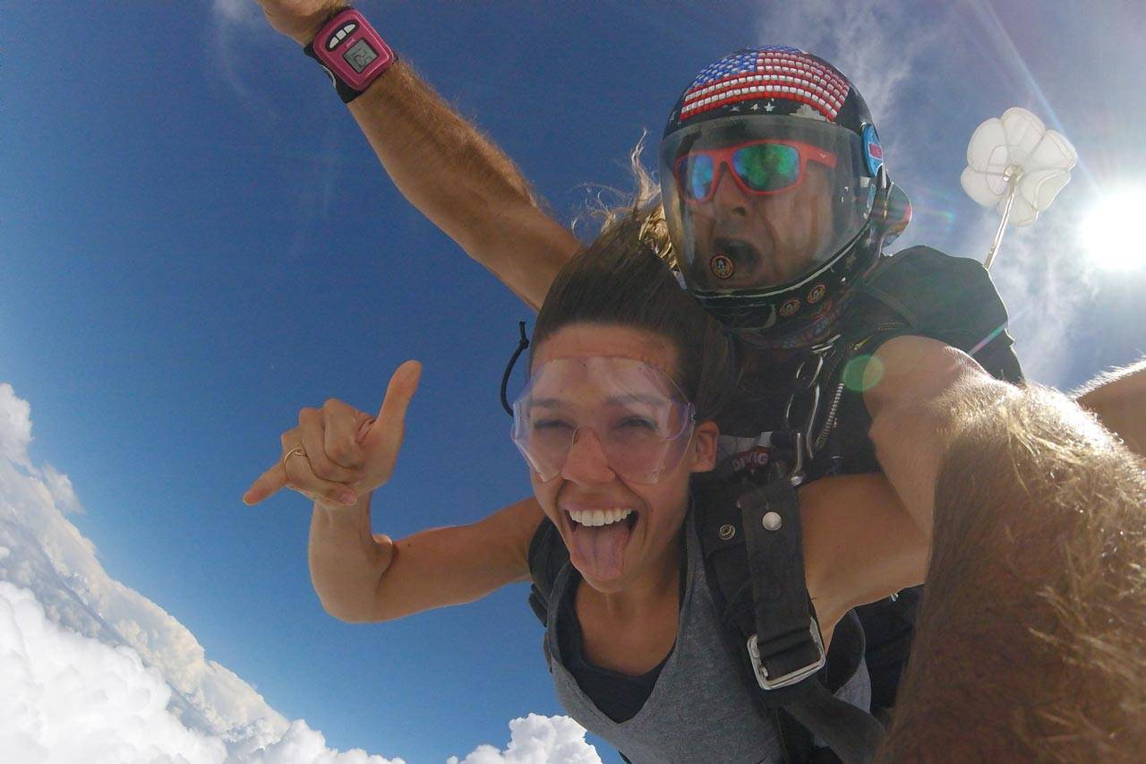 Women in gray shirt smiles with tongue out while in free fall with the skydiving company tandem instructor